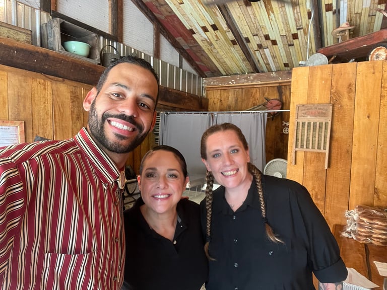 Three smiling people posing together inside a rustic wooden kitchen with exposed beams and natural lighting