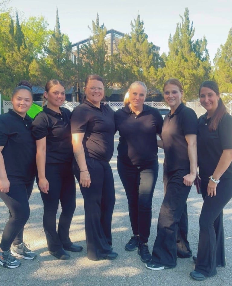 Six people in black uniforms standing together outdoors with cypress trees and industrial structure in the background
